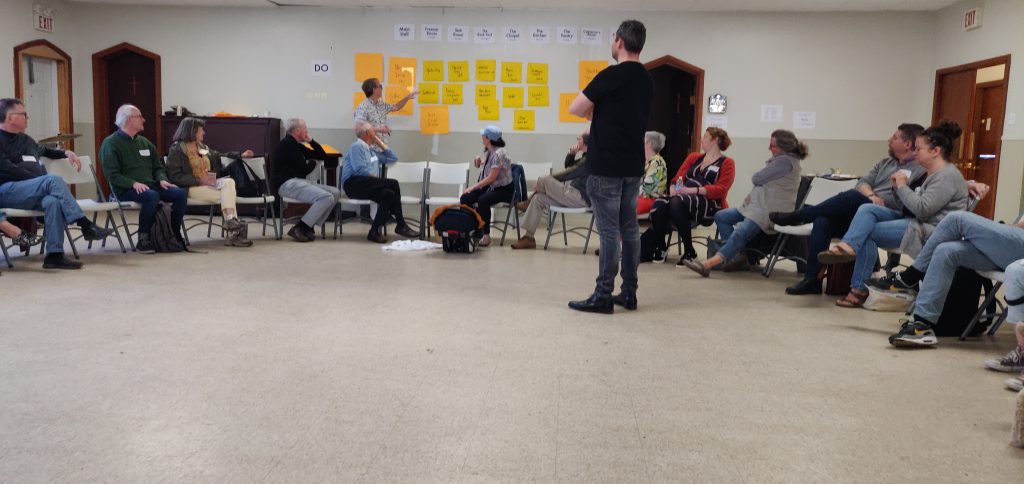 People sitting in a circle, looking at Peter Rukavina organising a wall-mounted conference schedule in real-time. Ton Zylstra facilitates.